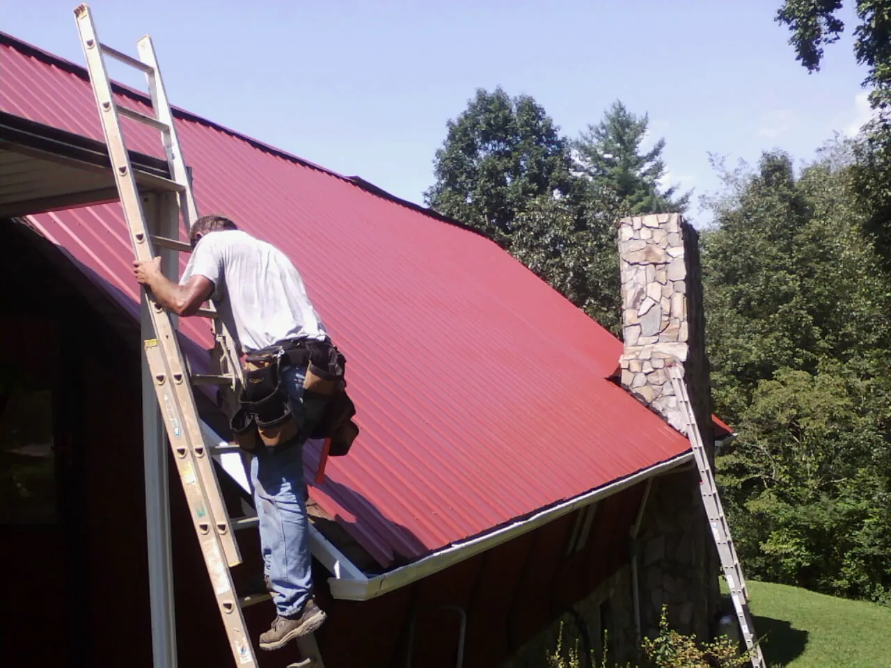 Roofer on a ladder beside a red metal roof and stone chimney — Soffit & Fascia Repair by Roofing Brothers in Scenery Hill